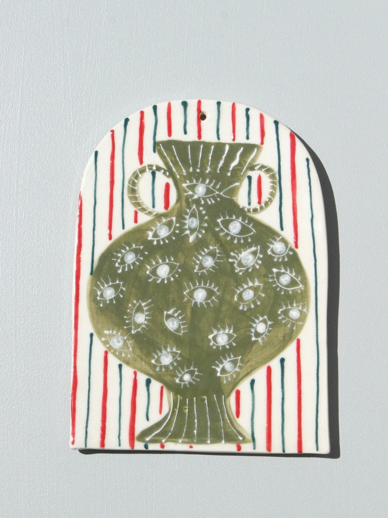 A handmade ceramic art tile featuring a green vase with a floral pattern, set against a striped blue and white background, with red accents.