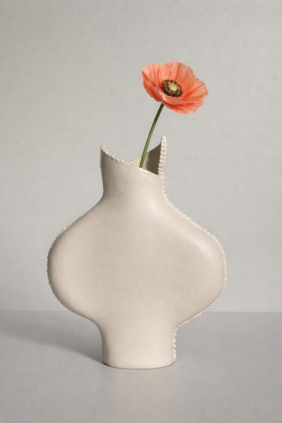 White ceramic vase with a unique design holding a single orange flower on a gray background