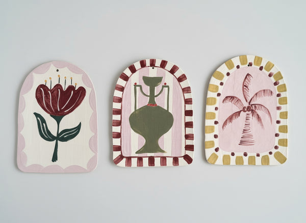 Three decorative wall hangings with floral, hourglass, and palm tree designs on a gray background.