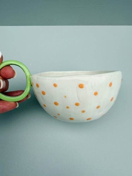 White ceramic cup with orange polka dots held by a hand against a light gray background