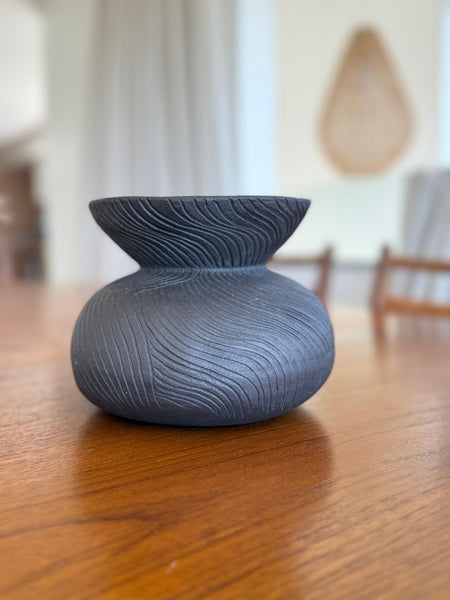 Black textured vase on a wooden table with a blurred indoor background